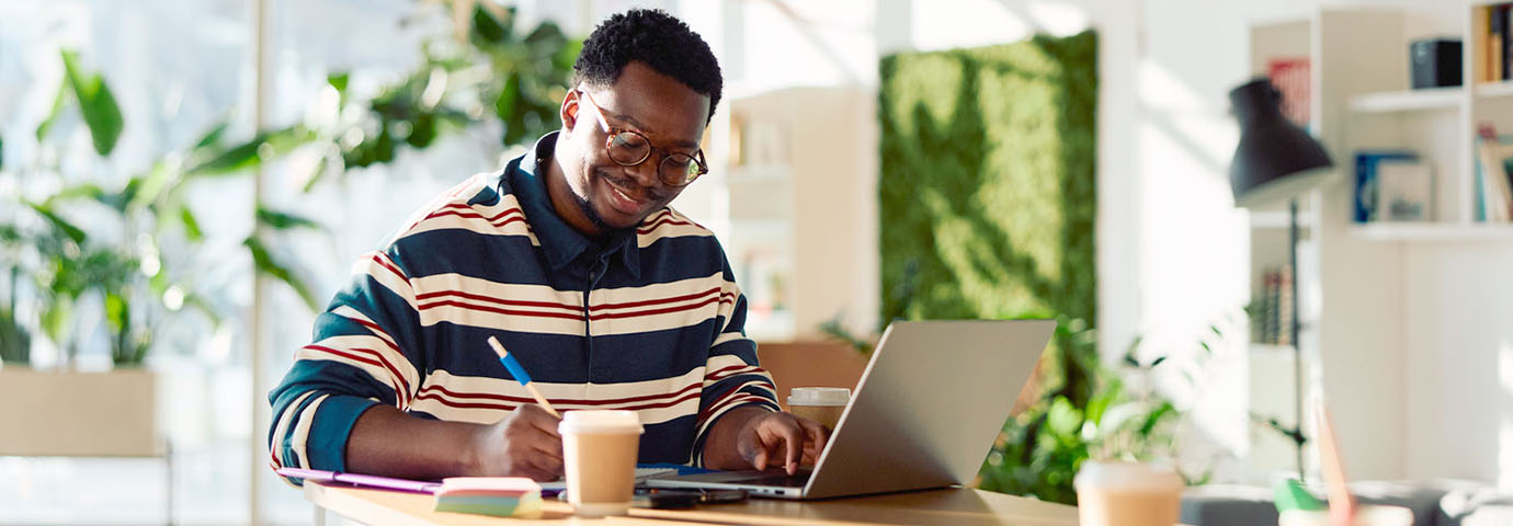 smiling student seated at desk writing while having one hand on laptop keyboard on table
